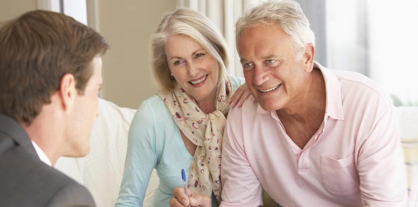 A young adult male in a suit with brown hair is looking at an older couple with white hair. The couple is has smiles on their faces. On the left is the wife. She is wearing a tan scarf with colorful spots and a long-sleeve teal shirt. To her right is her husband. He is wearing a light pink polo-style shirt.
