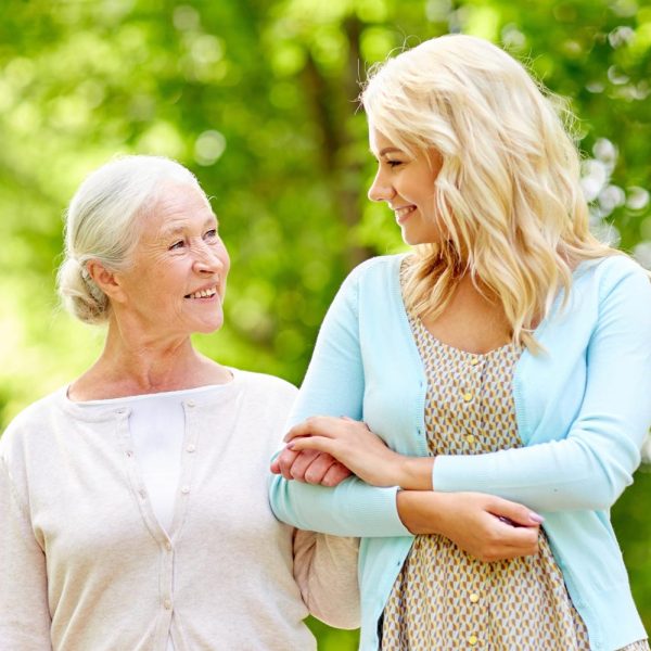 Two females are on a walk. On the left is an older female with white hair in a low bun, wearing a white shirt and cream cardigan. On her left is a blonde-haired younger female wearing a blue cardigan and a patterned neutral-colored shirt. Behind them are beautiful green trees.