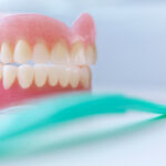 Dentures sitting on a white table with a teal A pair of dentures are sitting on a white table with a teal toothbrush in front of them.